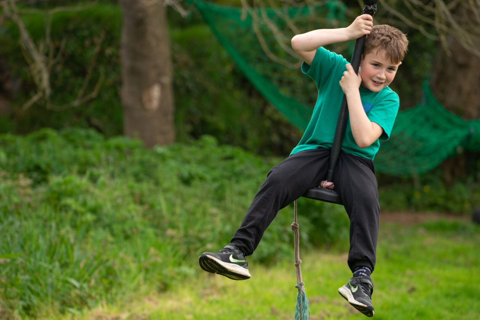 A young boy in a green t-shirt riding on a zip wire past green bushes and trees.